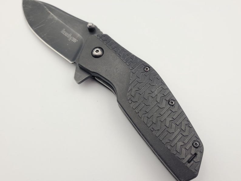 Op de afbeelding: Een zwart vouwmes met een gedeeltelijk gekarteld lemmet. Het mes heeft een getextureerde handgreep met een herhalend patroon en een zichtbaar draaipunt. Het lemmet is gemarkeerd met de merknaam "Kershaw".