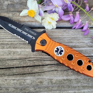 Puede incluir: Una navaja plegable TAC-FORCE naranja y negra con una hoja dentada y un símbolo médico. La navaja tiene un mango texturizado con varios recortes circulares y un pequeño gancho. La hoja es negra con el texto "TAC-FORCE".