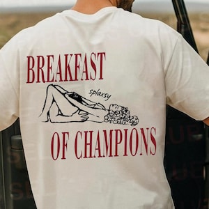 Puede incluir: Camiseta color crema con el texto "BREAKFAST OF CHAMPIONS" en borgoña. La camiseta presenta una ilustración en negro de una persona reclinada con un ramo de flores. La persona está de pie junto a un coche.
