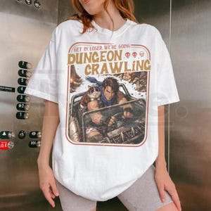 May include: White t-shirt with a graphic featuring a man driving a vehicle with a cat, and the text "GET IN LOSER. WE'RE GOING DUNGEON CRAWLING" in a brown frame. The design has a cartoonish style with a fantasy theme.