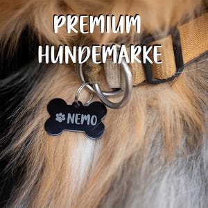 Può includere: Targhetta per cani a forma di osso nera con il nome "NEMO" in bianco e un disegno di impronta di zampa. La targhetta è appesa a un anello d'argento attaccato a un collare marrone. Il testo "PREMIUM HUNDEMARKE" è sopra.