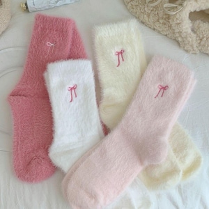 May include: Four pairs of fluffy socks in pink, white, and cream. Each sock features a small embroidered bow. The socks are soft and cosy, ideal for relaxing at home.