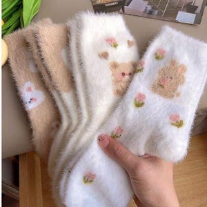 以下が含まれることがあります： ベージュと白の色合いのふわふわの靴下のコレクション。可愛らしい漫画の動物のデザインが特徴です。靴下はウサギ、クマ、花のアクセントを示しています。柔らかく、質感のある素材は、カジュアルウェアに最適な、居心地の良い快適な感触を提案しています。