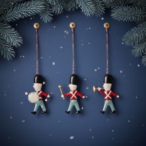 Op de afbeelding: Drie kerstversieringen in de vorm van speelgoedsoldaten die tegen een donkerblauwe achtergrond hangen. Elke soldaat draagt een zwarte hoed, een rode jas en een lichtblauwe broek. Eén houdt een trommel vast, één een baton en één een trompet. Bovenop liggen dennentakken.