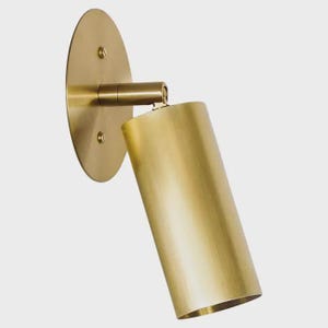 May include: A brass-colored wall-mounted spotlight fixture. The fixture has a circular base with three screws, a horizontal arm, and a cylindrical light head. The light head is angled downward.
