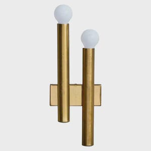 Puede incluir: Aplique de pared dorado con dos brazos cilíndricos verticales, cada uno coronado con una bombilla globo blanca. El aplique tiene una base rectangular y un diseño minimalista, adecuado para la iluminación interior.