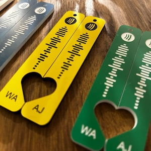 Op de afbeelding: Een verzameling kleurrijke, rechthoekige sleutelhangers met een hartvormige uitsparing. Elke sleutelhanger heeft een uniek geluidsgolfontwerp en de initialen "WA" en "AJ". De sleutelhangers zijn geel, groen, blauw en rood.