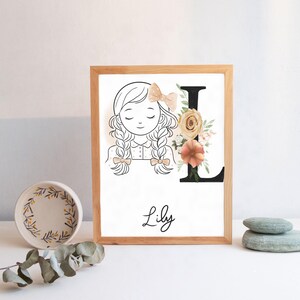 Könnte beinhalten: Holzgerahmter Kunstdruck mit einer Strichzeichnung eines Mädchens mit Zöpfen und einer Schleife. Der Buchstabe "L" ist mit Blumen verziert. Der Name "Lily" ist darunter in Schreibschrift geschrieben.