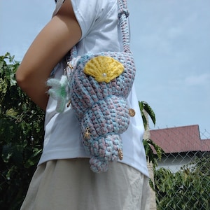 May include: A crochet crossbody bag shaped like a conch shell, crafted from variegated light blue and pink yarn. The bag features a yellow crocheted shell appliqué with a small white bead, and is decorated with tiny gold-toned starfish charms.