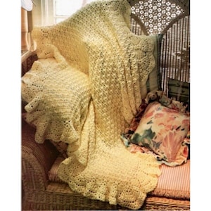 May include: A crocheted throw blanket in a pale yellow color with a ruffled edge. The blanket is draped over a wicker chair with a floral patterned pillow.
