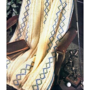 Op de afbeelding: Crèmekleurige gebreide deken met een ruitpatroon in blauw en groen. De deken is over een bruine leren fauteuil gedrapeerd. Een golfclub, een golfbal en een bronzen golfbeeld zijn zichtbaar.