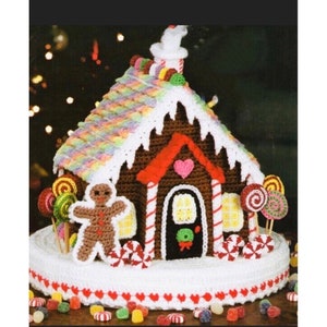 Op de afbeelding: Een gehaakt peperkoekhuisje met een bruin lichaam, een regenboogkleurig dak en witte randen. Het huis heeft een rood hart, een peperkoekmannetje en snoepdecoraties. De basis is wit met rode hartaccenten en kleurrijke snoepjes zijn verspreid.