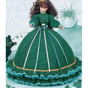 May include: A green crocheted doll dress with a white ribbon and a gold trim. The dress has a full skirt and a fitted bodice. The doll is wearing a white lace collar.