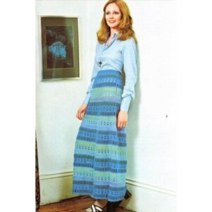 May include: A woman wearing a light blue long-sleeved shirt and a long, blue, green, and purple crocheted skirt. The skirt has a striped pattern and is made with open stitches.