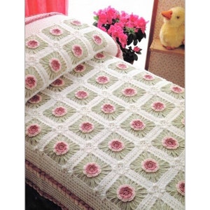 May include: A white and green crocheted blanket and pillow set with a floral pattern. The blanket features a diamond grid design with pink flower accents. The set is displayed on a bed, showcasing the handmade craftsmanship.