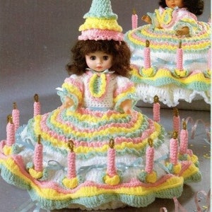 May include: Crochet pattern for a birthday cake dress and hat for a 13 inch music box doll or a 10 1/2 inch pillow doll. The dress is white with pink, yellow, and green ruffles. The hat is layered with pink, yellow, and green.