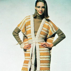 May include: A woman wearing a long, open-front crochet cardigan with horizontal stripes in yellow and white. The cardigan has a matching belt and fringe trim. She is also wearing a brown turtleneck, gray pants, and brown boots.