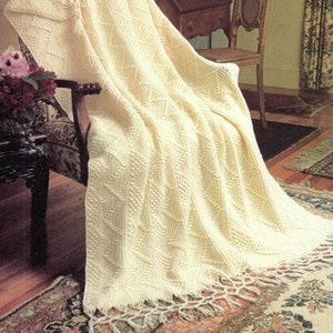 May include: A cream-colored knitted throw blanket draped over a chair. The blanket features a textured pattern with a fringed edge. The chair has a patterned fabric seat and back. The room has a vintage aesthetic.