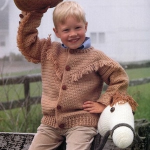 May include: A young boy wearing a brown crocheted cardigan with fringe trim and a brown cowboy hat. He is holding a white crocheted toy horse.