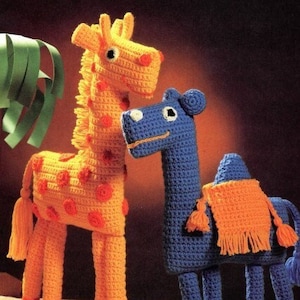 May include: A crocheted giraffe with orange and red spots stands next to a crocheted camel with a blue body and orange blanket. Both stuffed animals have large, black eyes.