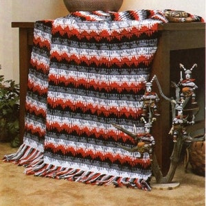 May include: A crocheted throw blanket with a striped pattern in red, white, black, and gray. The blanket has a fringe border and is draped over a wooden table.