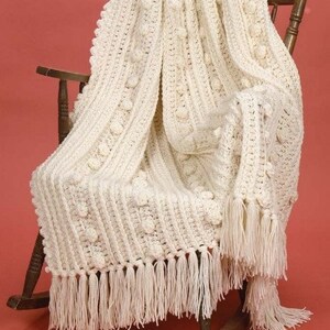 May include: A white crocheted throw blanket with a fringe border and a textured bobble stitch pattern. The blanket is draped over a wooden rocking chair.