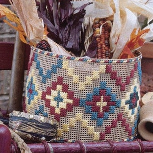 Può includere: Un cesto cilindrico intrecciato con un motivo geometrico rosso, blu e giallo. Il cesto è riempito con mais secco, foglie e altri elementi naturali, creando un'esposizione autunnale rustica.