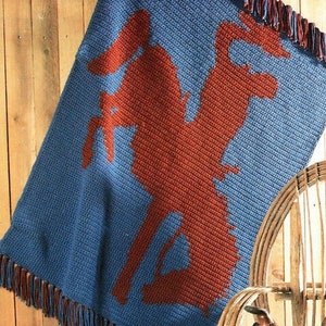 Puede incluir: Una manta de crochet con borde de flecos. La manta es azul con una silueta roja de un vaquero montando a caballo.