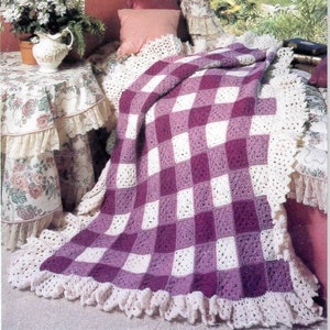 Op de afbeelding: Een gehaakt deken met een wit en paars ruitpatroon. Het deken heeft een witte gehaakte rand met een gerimpelde zoom.