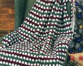 May include: A crocheted throw blanket with a striped pattern in white, green, and burgundy. The blanket is draped over a chair and is near a Christmas tree.