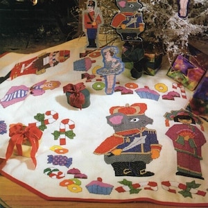May include: A white Christmas tree skirt with a red border, featuring embroidered images of a nutcracker, a ballerina, a candy cane, and a mouse wearing a crown and holding a sword. The skirt is decorated with colorful candies, cupcakes, and donuts.