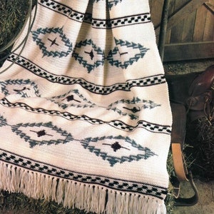 Könnte beinhalten: Eine weiße Häkeldecke mit einem geometrischen Muster in Blautönen, Schwarz und Braun. Die Decke hat einen Fransenrand und ist über einen Heuballen gelegt.