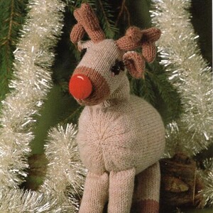 May include: A knitted reindeer figurine with a red nose, brown antlers, and black eyes. The reindeer is beige with brown accents on the face and legs. The background includes green pine needles and silver tinsel, suggesting a holiday theme.