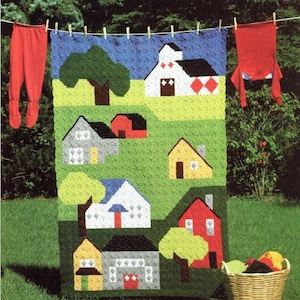 Puede incluir: Una manta de ganchillo que representa una escena campestre colorida con casas, árboles y un cielo azul. La manta está hecha con hilo de diferentes colores y presenta una variedad de puntadas.