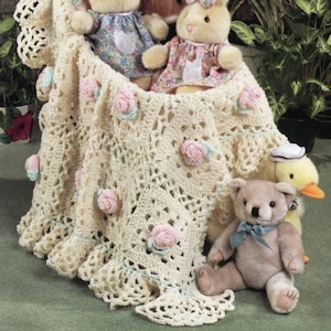May include: A crocheted baby blanket with a lacy border and pink flower accents. The blanket is draped over a group of stuffed animals, including a teddy bear, a duck, and three bunnies.