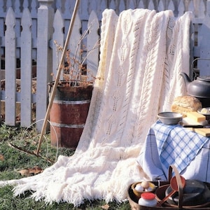 May include: A white knitted throw blanket with a lacy pattern, draped over a wooden barrel and a picnic table with a checkered tablecloth. The blanket has fringe along the edges.