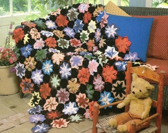Op de afbeelding: Een handgemaakte gehaakte deken, gedrapeerd over een bank. De deken heeft een zwarte achtergrond met verschillende bloemvormen in tinten rood, blauw, roze en wit. Een kleine teddybeer zit op een miniatuur houten stoel.