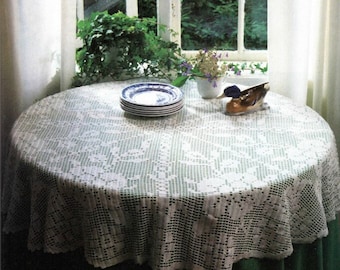 May include: A round table covered with a white, open-weave lace tablecloth. A stack of blue and white plates sits on the table, along with a decorative duck figurine and a small vase of flowers. The table is near a window.
