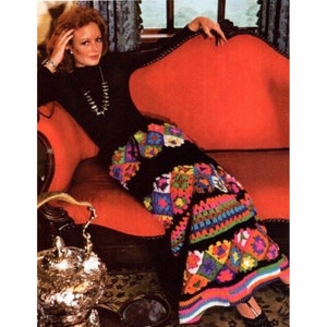 May include: A woman wearing a black turtleneck sweater and a long, colourful crochet skirt. The skirt is made of granny squares in various colours, including pink, green, blue, yellow, and orange. The skirt is accented with black trim and a black belt.