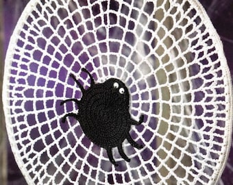 Può includere: Acchiappasogni in crochet bianco a forma di ragnatela con un ragno in crochet nero al centro.