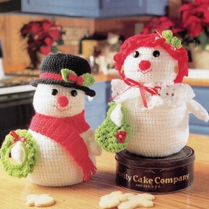 May include: Two crocheted snowmen. One snowman wears a black hat, red scarf, and holds a green wreath. The other snowman has red hair, a white dress, and a green purse. Both have black eyes, red noses, and smiles. The snowmen are on a wooden surface.