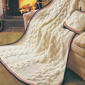 May include: Cream-colored cable knit blanket and matching pillow with a black and red border. The blanket is draped over a beige sofa, with a fireplace in the background. The cozy, textured design is visible.