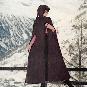 May include: A long, dark purple knitted cape with a hood and a tassel. The cape is worn by a person standing in a snowy landscape.