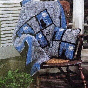 May include: A crocheted throw blanket and pillow with a blue and gray color scheme. The blanket and pillow feature a repeating pattern of black cats and white dandelions. The throw is draped over a wooden chair.
