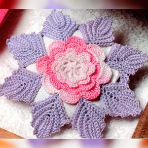May include: A crocheted flower motif in shades of pink and purple. The flower is surrounded by eight crocheted leaves in a starburst pattern.