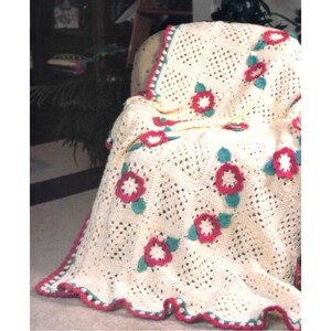 May include: A crocheted throw blanket with a white background and a repeating pattern of pink and green flowers. The blanket is draped over a chair.