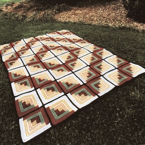 May include: A crocheted blanket with a log cabin pattern in shades of brown, green, white, and beige. The blanket is laid out on a grassy surface.