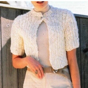 May include: Cream-colored, short-sleeved knitted bolero sweater. The sweater has a round neckline and a single button closure. The model is wearing a beige belt and pants. The background is a wooden fence.