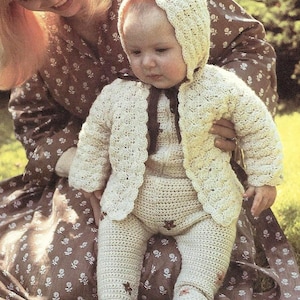 May include: A woman in a brown floral dress holds a baby wearing a crocheted white outfit with a bonnet. The baby is sitting on the woman's lap and looking at the camera.