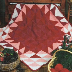Peut inclure: Une couverture crochetée à la main, rouge, blanche et rose, avec un motif géométrique en forme d'éclat d'étoile. La couverture est exposée sur une surface en bois, avec des paniers de cadeaux et de sphères rouges à proximité. Les dimensions de la couverture ne sont pas visibles.